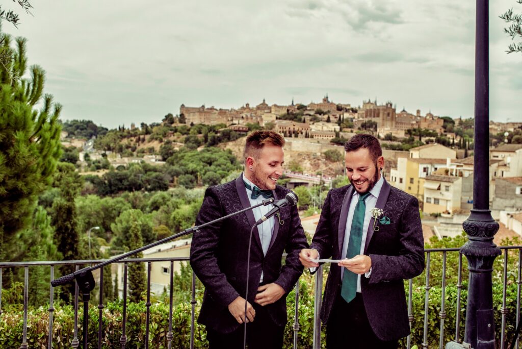 Two men in suits share a joyful moment with a scenic city backdrop during their outdoor wedding ceremony.