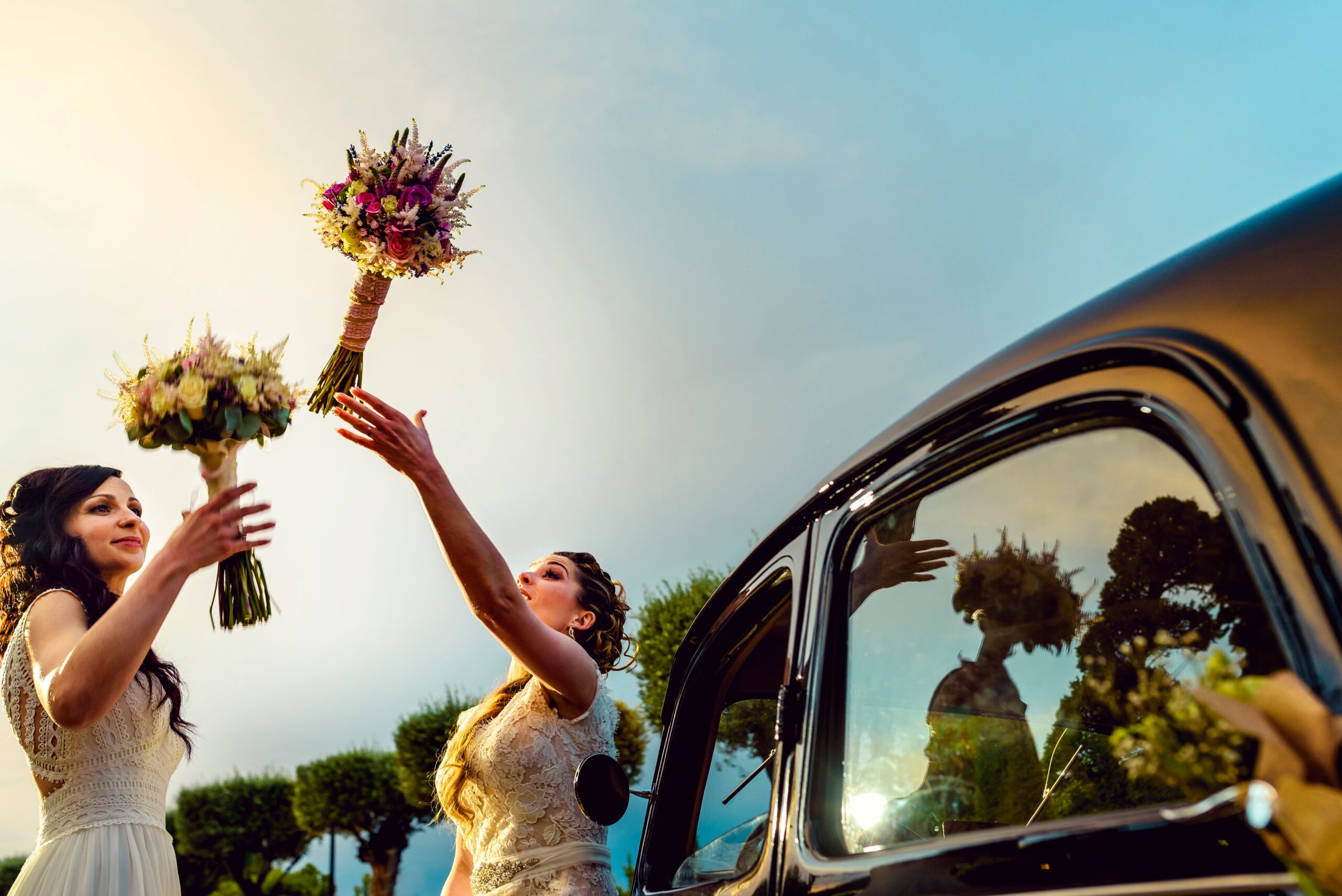 Two brides joyfully exchange bouquets beside a vintage car at their vibrant wedding in Toledo.